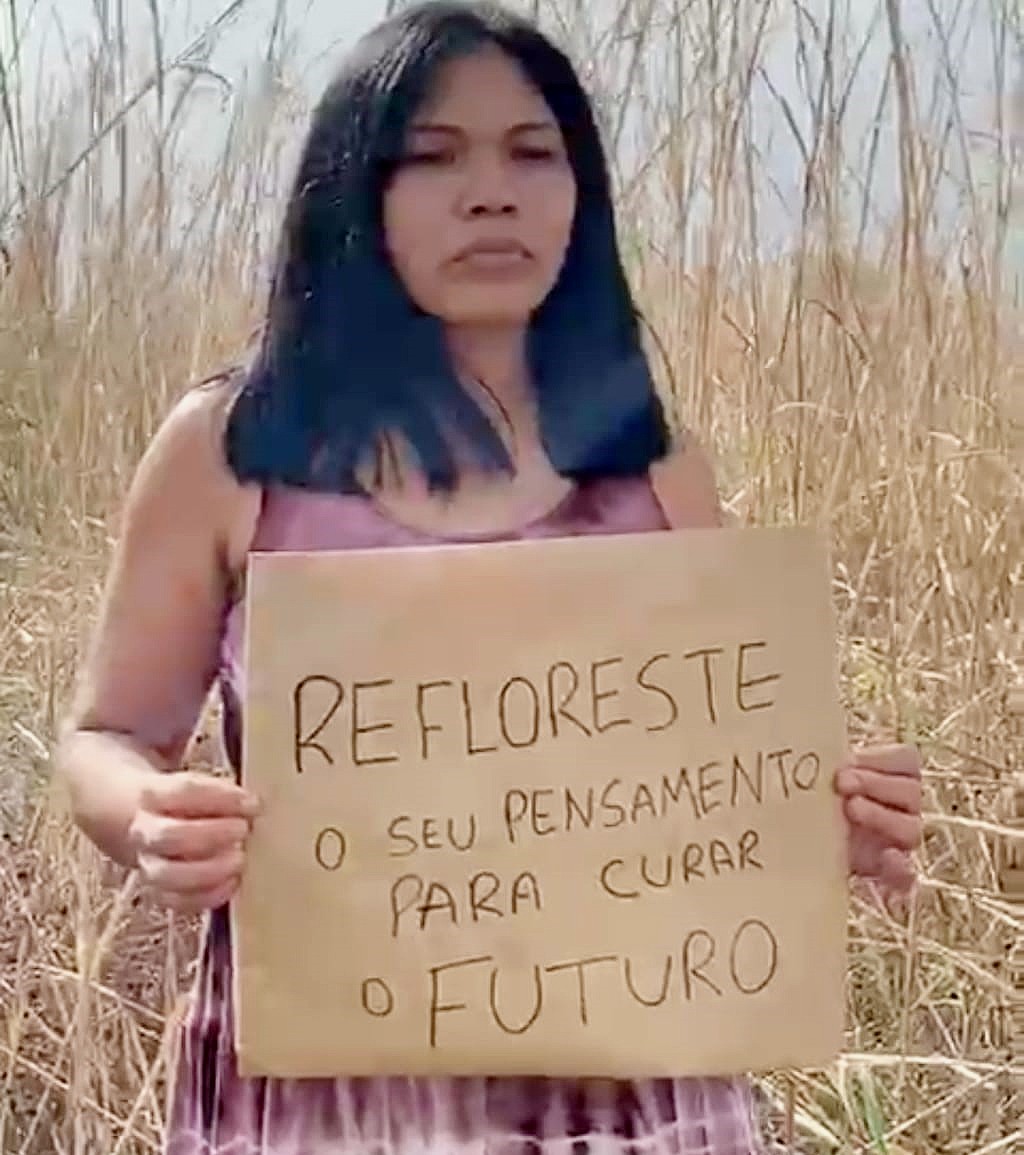 Indigenous woman holding a sign REFLORESTE o seu pensamento para curar o futuro - reforest your thinking to heal the future